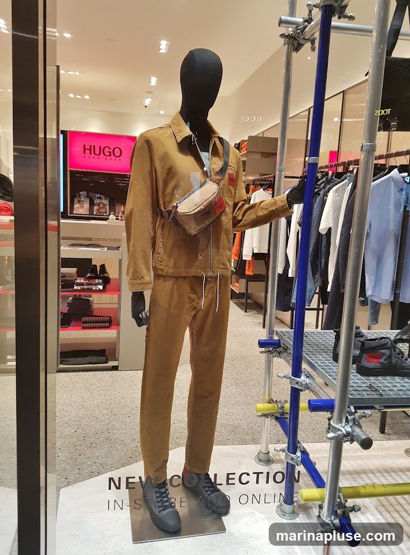 A mannequin dressed in a tan jacket and pants, wearing a crossbody bag, stands in a store window. Behind the mannequin, racks of clothing are visible, along with a large pink sign that reads "HUGO HUGO BOSS". Text on the floor reads "NEW COLLECTION IN-STORE AND ONLINE".