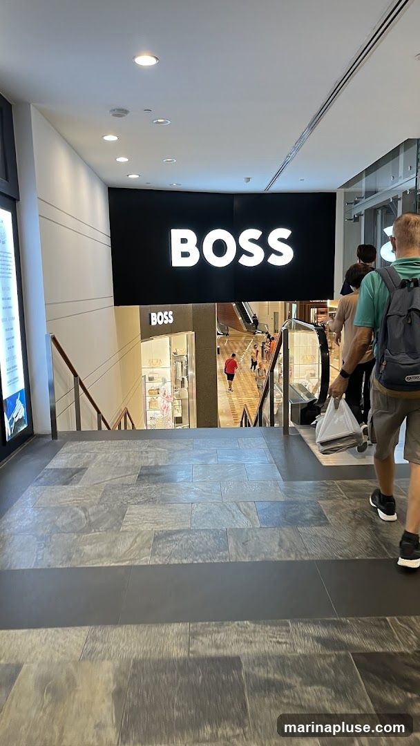 A view looking down a set of stairs towards a BOSS store entrance. A large black sign with the white BOSS logo is visible above the store. Inside the store, mannequins display clothing and handbags. People are seen walking on escalators and in the background.