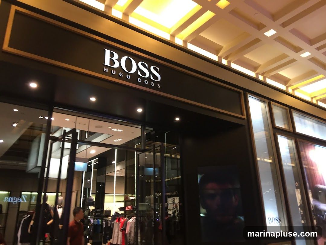 The exterior of a BOSS Hugo Boss store. The store has a dark facade with a prominent sign that reads "BOSS HUGO BOSS" in white lettering. Large glass windows display clothing and mannequins inside. The ceiling above the entrance is illuminated with a grid of lights.