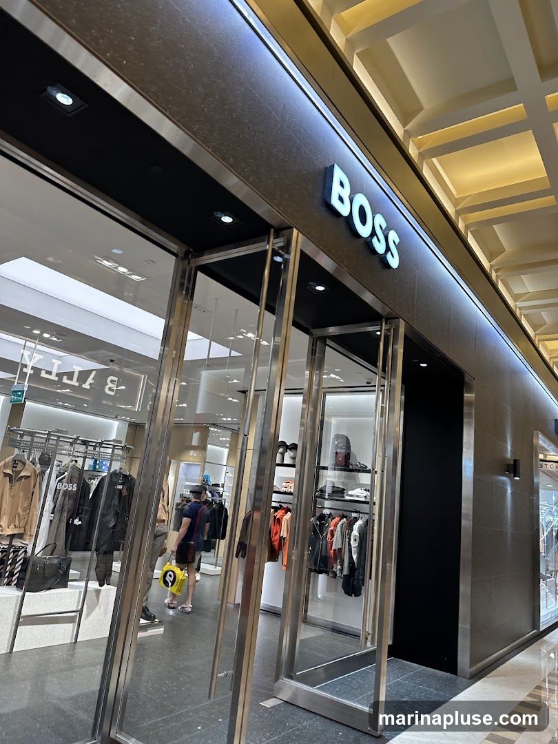 The exterior of a BOSS store with large glass doors and windows. The BOSS logo is prominently displayed in illuminated white lettering above the entrance. Inside, racks of clothing and accessories are visible, including jackets, shirts, bags, and shoes.