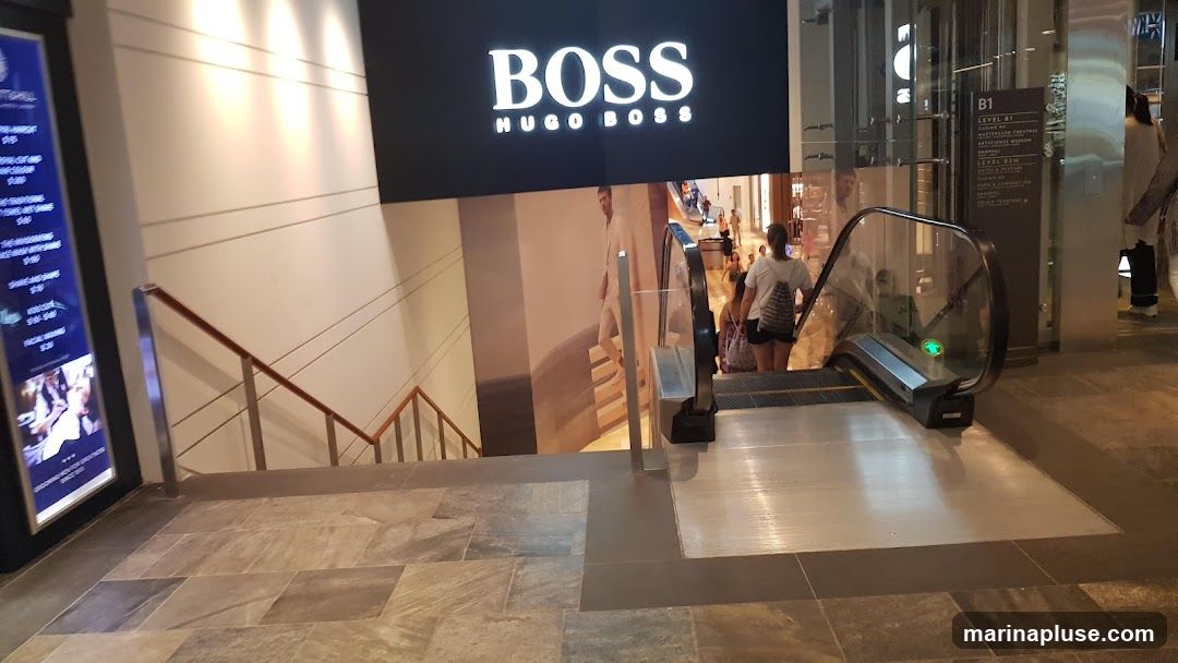 A large BOSS HUGO BOSS sign is displayed above an escalator. A large advertisement featuring a man in a suit is on the wall behind the escalator. To the left, a digital menu board displays various services and prices.