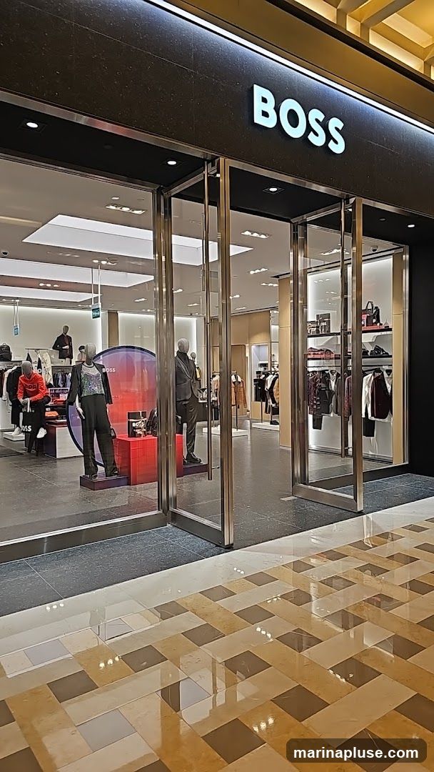 The exterior of a BOSS store with a prominent illuminated sign above the entrance. The storefront features large glass windows and doors, showcasing mannequins dressed in clothing and displays of merchandise inside. The floor in front of the store has a patterned tile design.