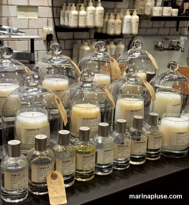 A display of various scented candles under glass domes and small perfume bottles arranged on a dark surface. The candles and bottles have labels with names like "SANTAL 26", "FIGUE 15", "CEDRE 11", and "PETIT GRAIN 21". In the background, shelves are stocked with rows of white bottles.