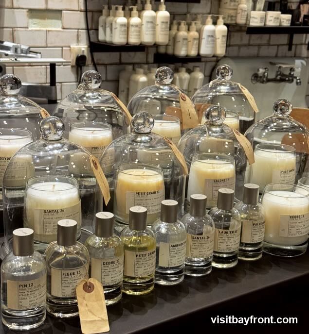 A display of various scented candles under glass domes and small perfume bottles arranged on a dark surface. The candles and bottles have labels with names like "SANTAL 26", "FIGUE 15", "CEDRE 11", and "PETIT GRAIN 21". In the background, shelves are stocked with rows of white bottles.