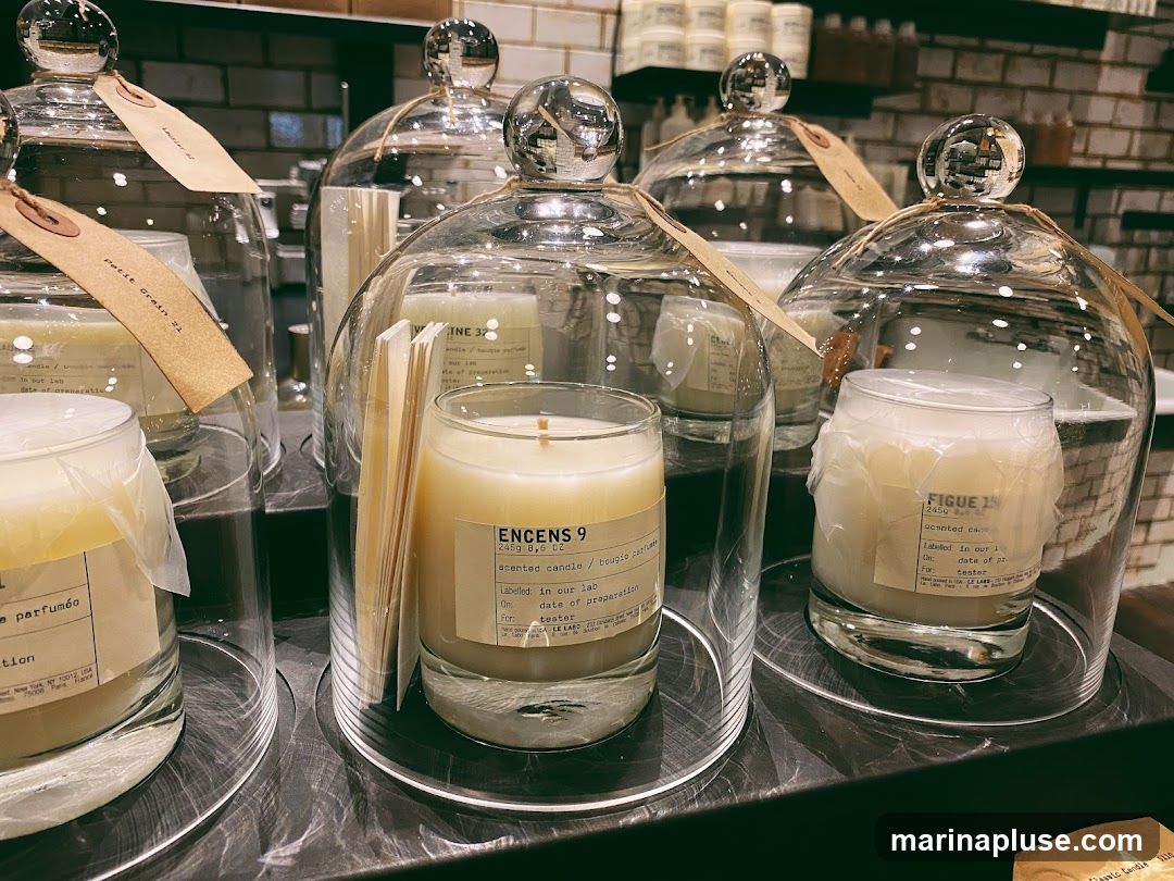 A display of several scented candles, each under a glass cloche. The candles are in glass jars and have labels with product information. Some labels are partially obscured.