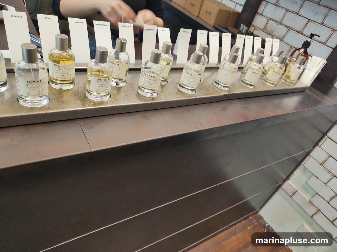 A display of various perfume bottles on a metal tray, with white cards indicating the names of the scents. The bottles are arranged in rows, and some have labels with text. In the background, a person's hands are visible, suggesting a retail environment. The counter is made of dark metal, and the wall behind it is tiled.
