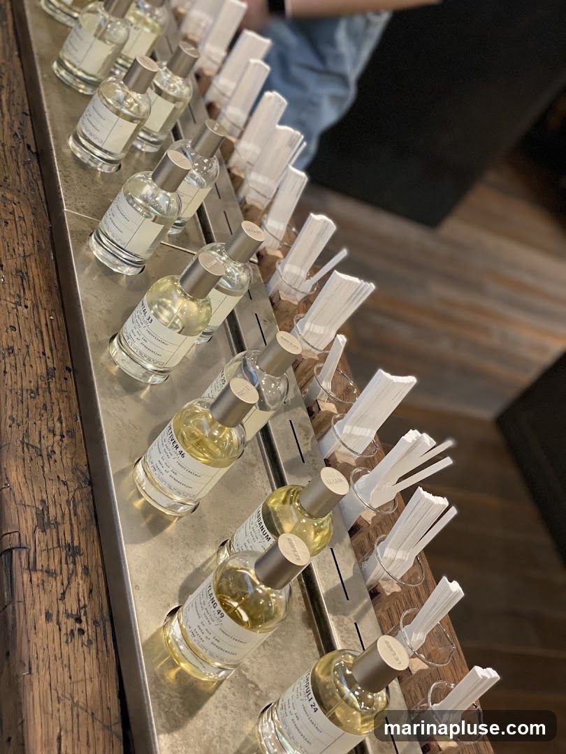 A close-up, angled shot showcases a collection of perfume bottles arranged on a metal display stand. The bottles are clear glass with silver caps, and each contains a pale yellow liquid. Labels on the bottles are visible, with some text readable, such as 'GYPSY WATER 33', 'TETIVER 46', 'YLANG 49', and 'TAMBULI 24'. Next to the perfume bottles are rows of white paper scent strips, each held upright in small glass or wooden holders. The background is blurred, suggesting an indoor retail environment with wooden flooring and dark elements. A person's arm and shoulder are visible in the upper right corner, wearing a light blue garment.