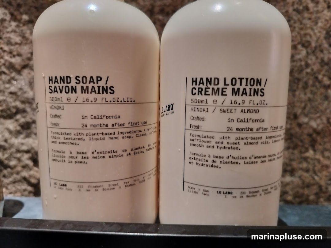 Two white pump bottles of hand soap and hand lotion. The labels are white with black text. The hand soap is HINOKI scented and the hand lotion is HINOKI / SWEET ALMOND scented. Both are 500ml / 16.9 FL.OZ. The bottles are placed on a dark shelf with a textured background.