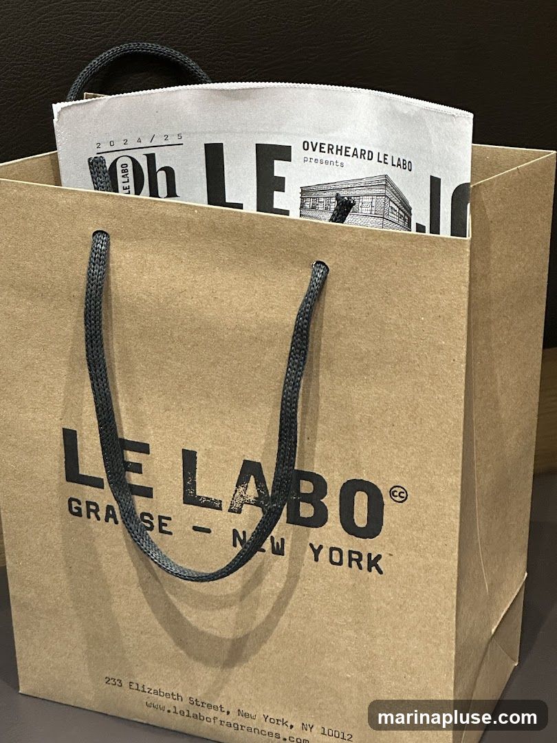 A brown paper shopping bag with dark gray handles. The bag features the Le Labo logo and text, including "GRASSE - NEW YORK" and the address "233 Elizabeth Street, New York, NY 10012" along with the website "www.lelabofragrances.com". A folded paper insert with "OVERHEARD LE LABO presents" and "2024/25" is visible inside the bag.
