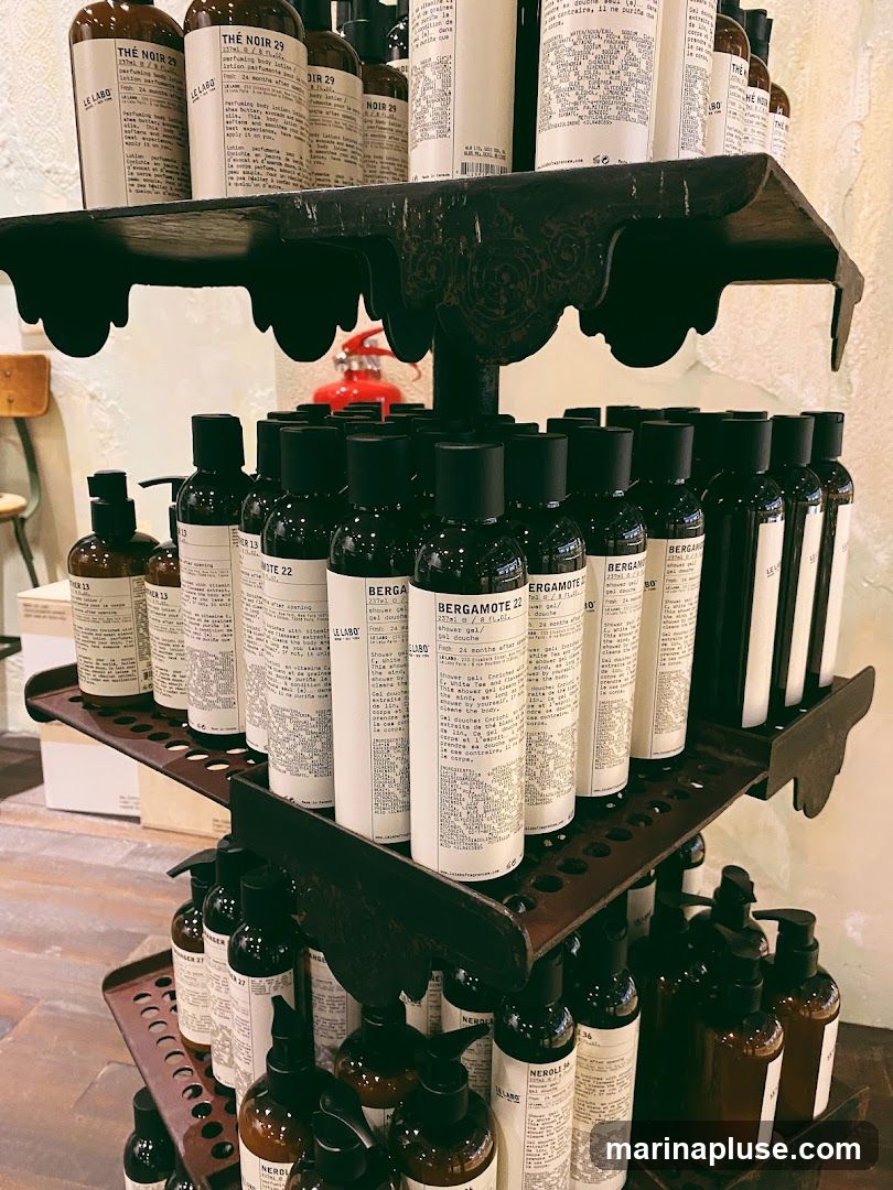 A multi-tiered display rack filled with various Le Labo branded products, including perfumes and body lotions. The bottles are predominantly dark glass with white labels featuring product names and descriptions. Some labels are partially visible, showing names like "THE NOIR 29", "BERGAMOTE 22", and "NEROLI 36". The display appears to be in a retail setting.