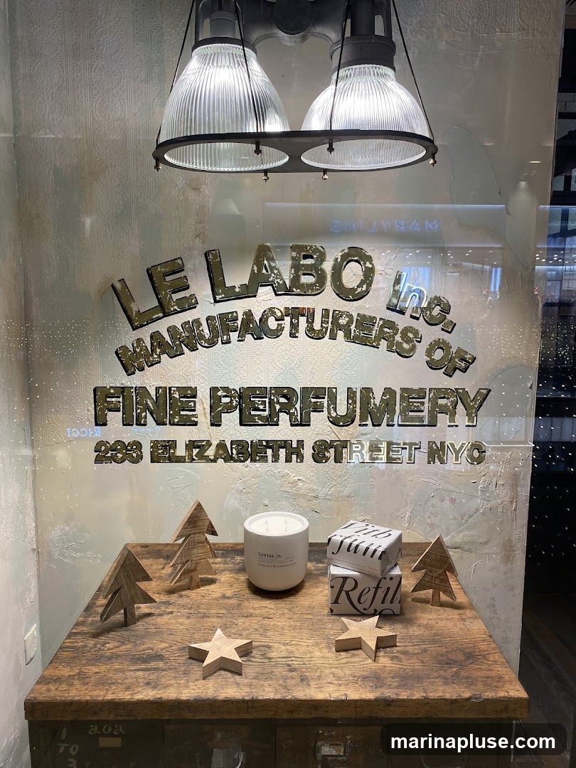 A window display for Le Labo, featuring the company name and address on the glass. The display includes a white candle labeled SANTAL 26, two boxes with the word "Refil" printed on them, and several wooden decorative items shaped like trees and stars. Two industrial-style lamps hang above the display.