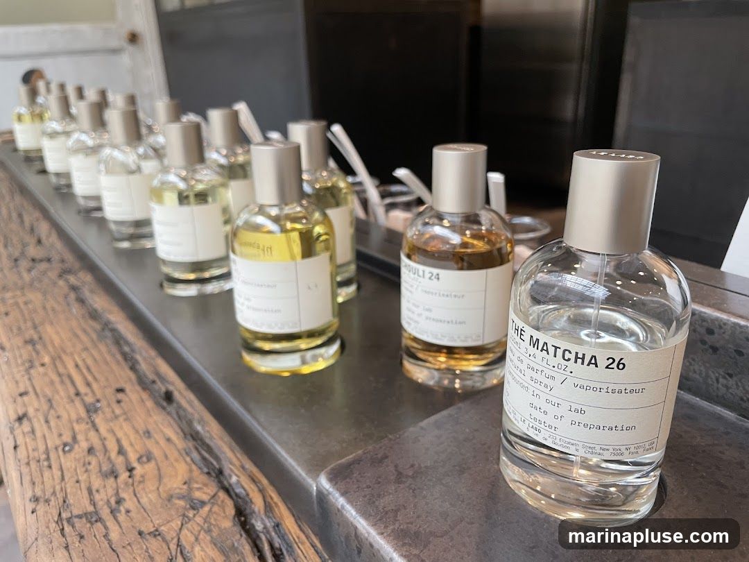 A display of perfume bottles on a wooden counter. The bottles are clear glass with silver caps and have white labels. One bottle in the foreground is labeled "THE MATCHA 26". Another bottle behind it is labeled "QUL1 24".