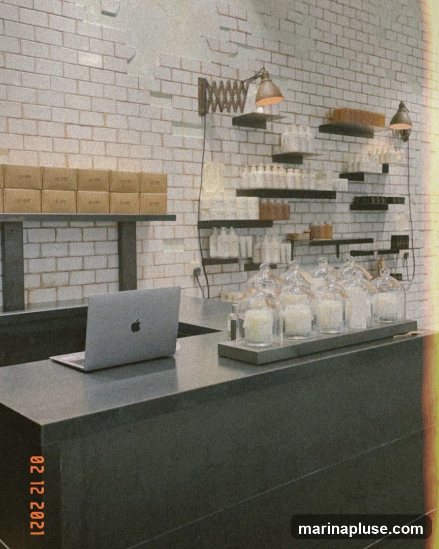 A dark counter with a laptop and several glass-covered candles is in the foreground. Behind the counter, shelves are stocked with bottles and boxes. The wall is tiled with white bricks.
