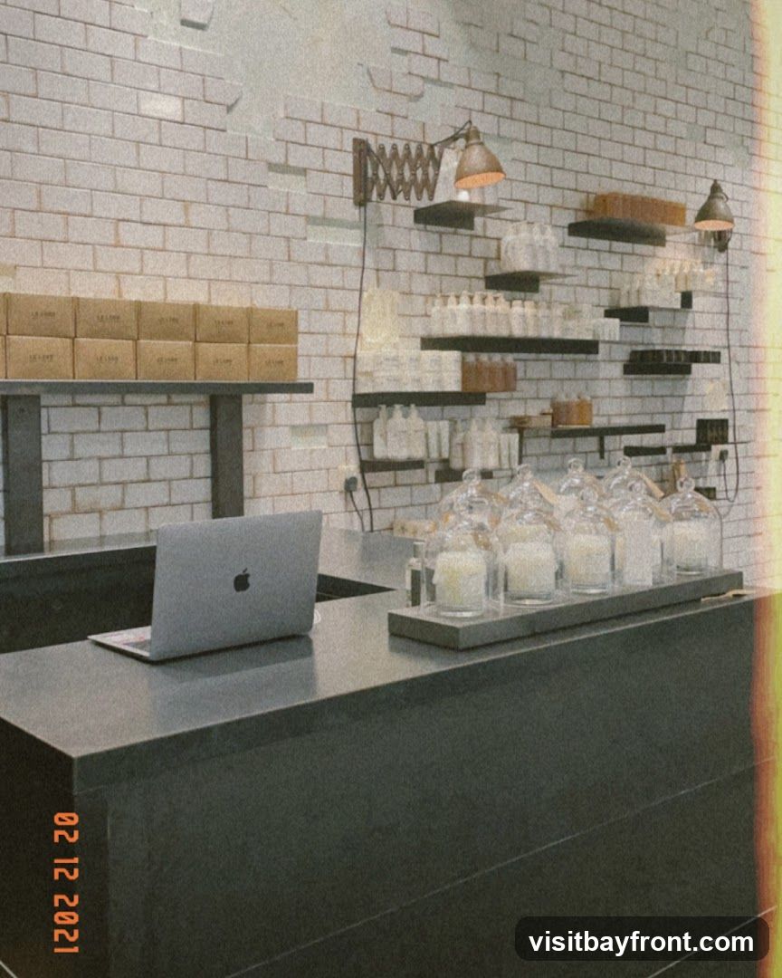 A dark counter with a laptop and several glass-covered candles is in the foreground. Behind the counter, shelves are stocked with bottles and boxes. The wall is tiled with white bricks.