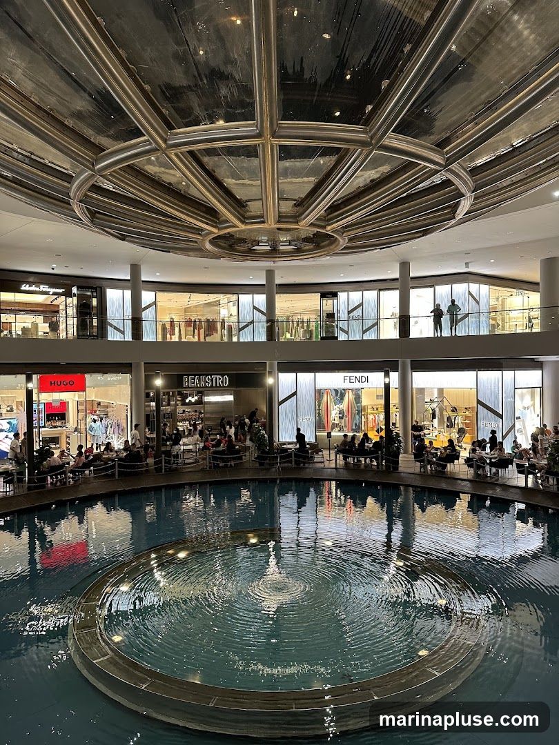 An indoor shopping mall features a large circular fountain in the center, with shops and restaurants surrounding it on multiple levels. The ceiling has a reflective, metallic design. Visible shop names include HUGO, BEANSTRO, and FENDI. People are seated at tables around the fountain and browsing in the shops.