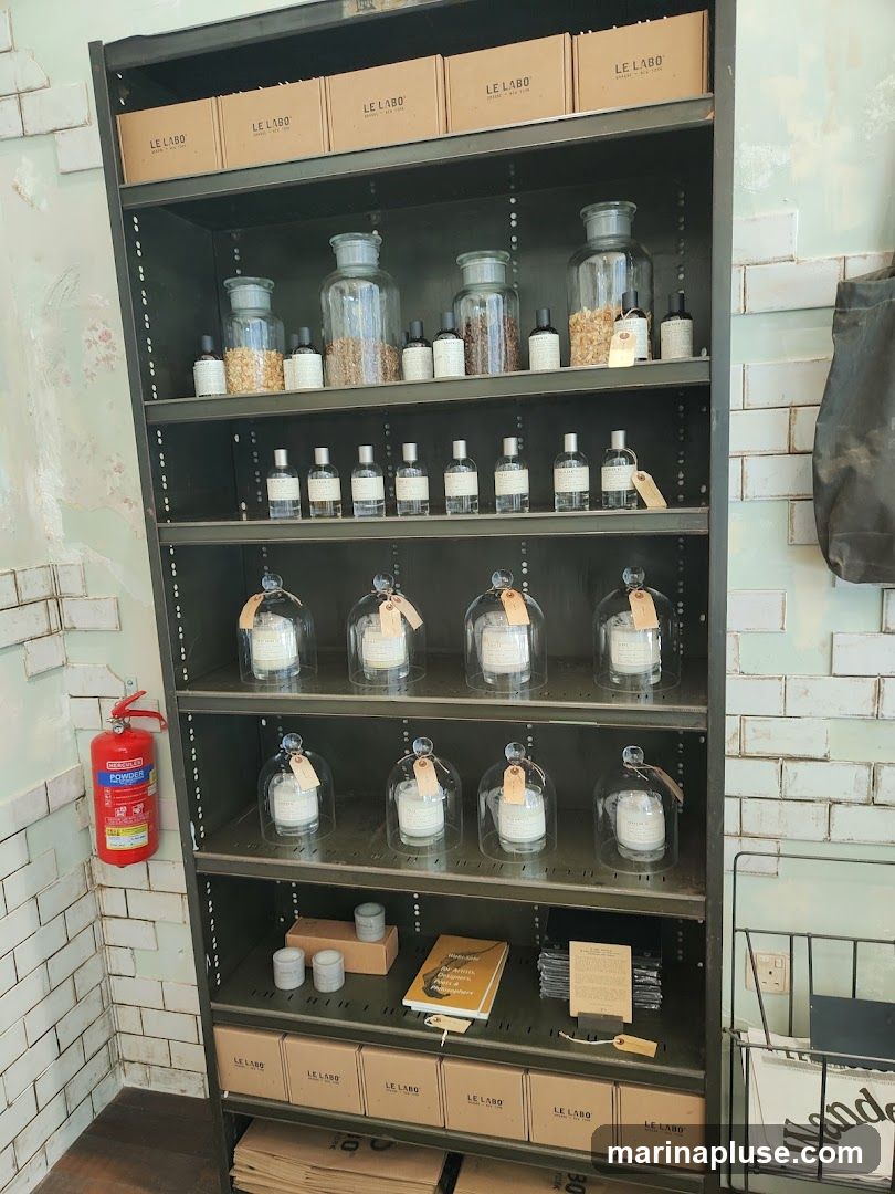 A tall, dark metal shelf unit displays various Le Labo products. The top shelf has several Le Labo branded boxes. Below that are large glass jars filled with what appear to be raw ingredients, alongside smaller bottles of fragrance. The next two shelves feature candles in glass domes. The shelf below that has small cylindrical items, a yellow book, and stacks of paper. The bottom shelves are filled with more Le Labo branded boxes. A red fire extinguisher is mounted on the wall to the left. The background features tiled walls with a distressed paint effect.