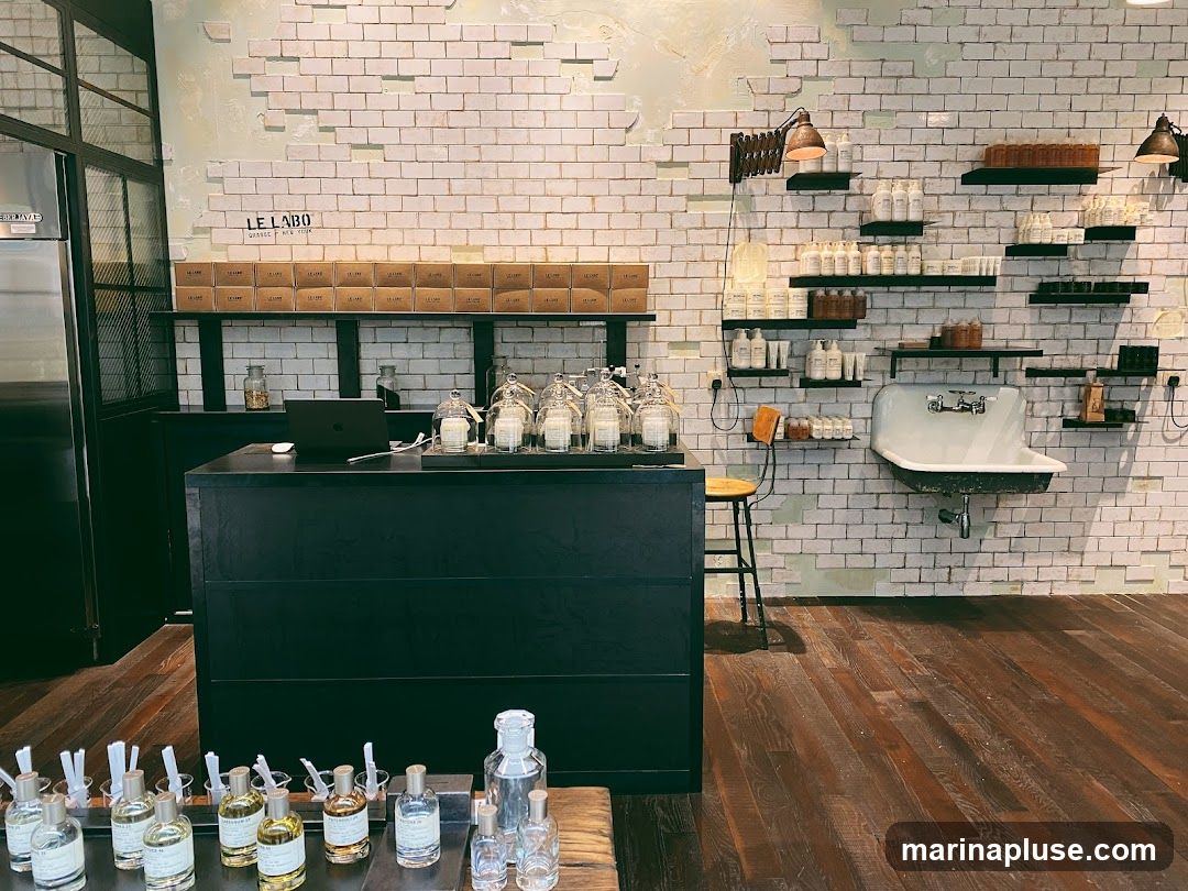 The interior of a Le Labo store with a black counter in the foreground displaying various perfume bottles. Behind the counter, shelves are stocked with products and boxes. The back wall is made of white brick and features more shelves with bottles and a sink. The floor is made of dark wood planks.