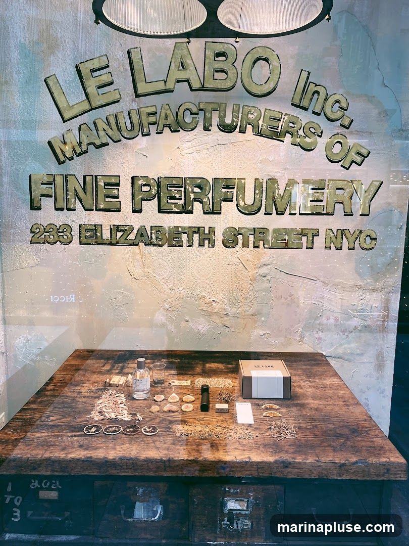 A display inside a Le Labo store, featuring a wooden table with various perfume ingredients, a bottle of perfume, and a Le Labo branded box. The store's name and address, "LE LABO Inc. MANUFACTURERS OF FINE PERFUMERY 233 ELIZABETH STREET NYC", are visible on the wall behind the table.