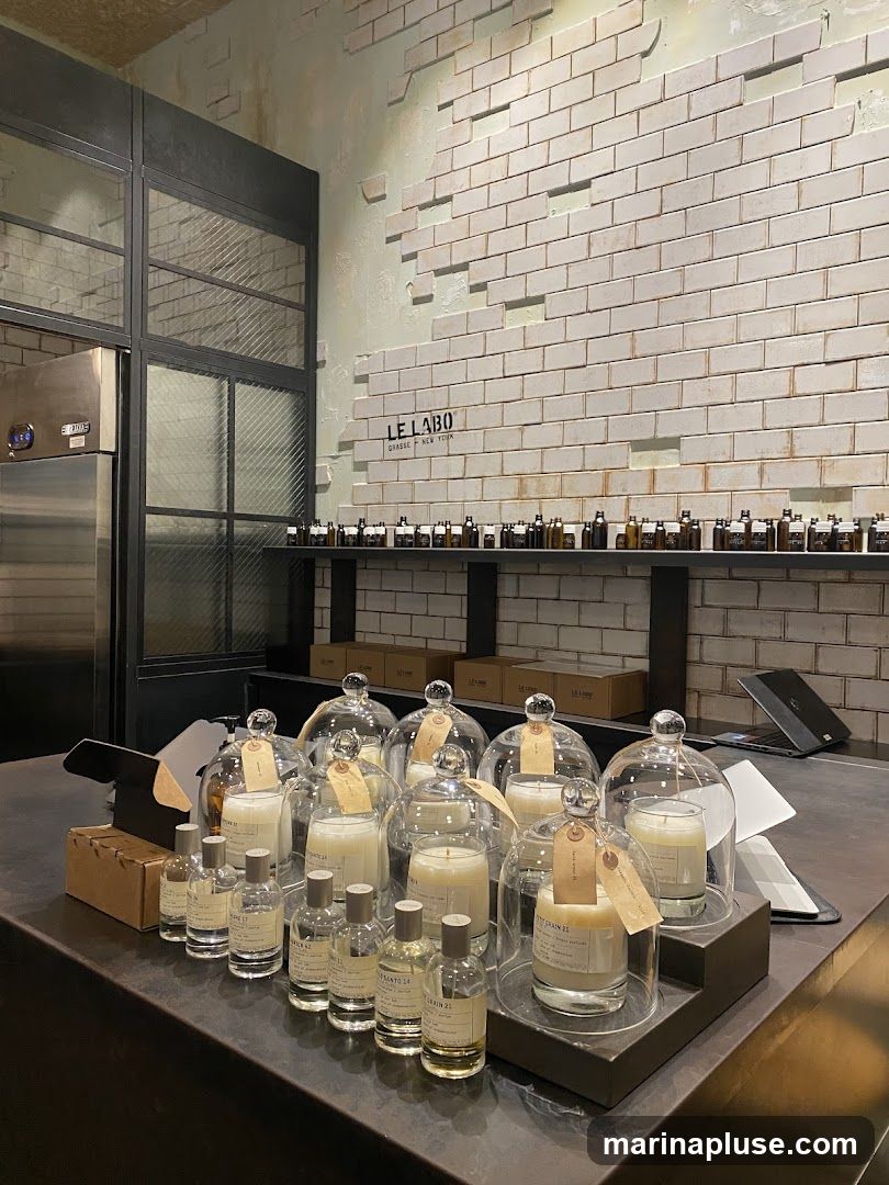 A display of candles and perfume bottles on a counter in a store. The wall behind the counter is tiled with white bricks and has the "LE LABO GRASSE NEW YORK" logo. Shelves above the logo are lined with small amber bottles.