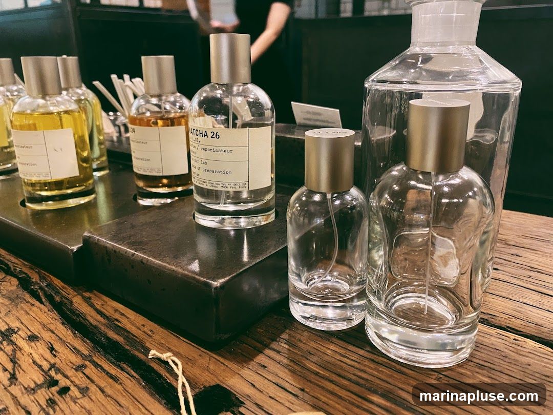 A close-up shot of several perfume bottles displayed on a rustic wooden surface. The bottles are made of clear glass with metallic caps. Some bottles contain yellow or amber-colored liquid, while others are empty. One bottle in the center has a label that reads "MATCHA 26" and "eau de parfum / vaporisateur". In the background, a person is partially visible, and there are more bottles and display elements.