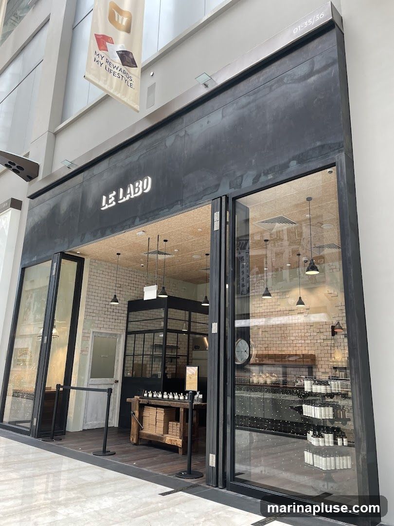 The exterior of a Le Labo store with large glass doors and windows. The store's name, "LE LABO," is displayed in illuminated white lettering above the entrance. Inside, shelves are stocked with bottles and boxes, and a wooden workbench is visible. A banner hangs above the store with the text "MY REWARDS. MY LIFESTYLE."