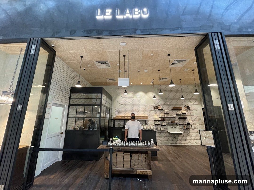 The exterior of a Le Labo store with open glass doors. A person wearing a mask stands behind a wooden counter displaying bottles. The store interior features white brick walls, shelves with products, and industrial-style lighting. The store name "LE LABO" is illuminated above the entrance.
