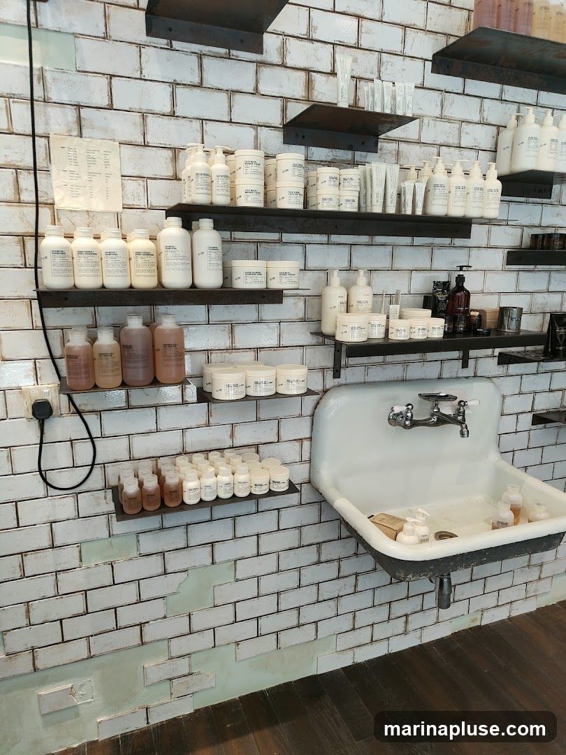Shelves on a white brick wall display various bottles and jars of hair and skin care products. A large white sink with a faucet is mounted on the wall to the right. The products are mostly in white or clear bottles with minimalist labeling.