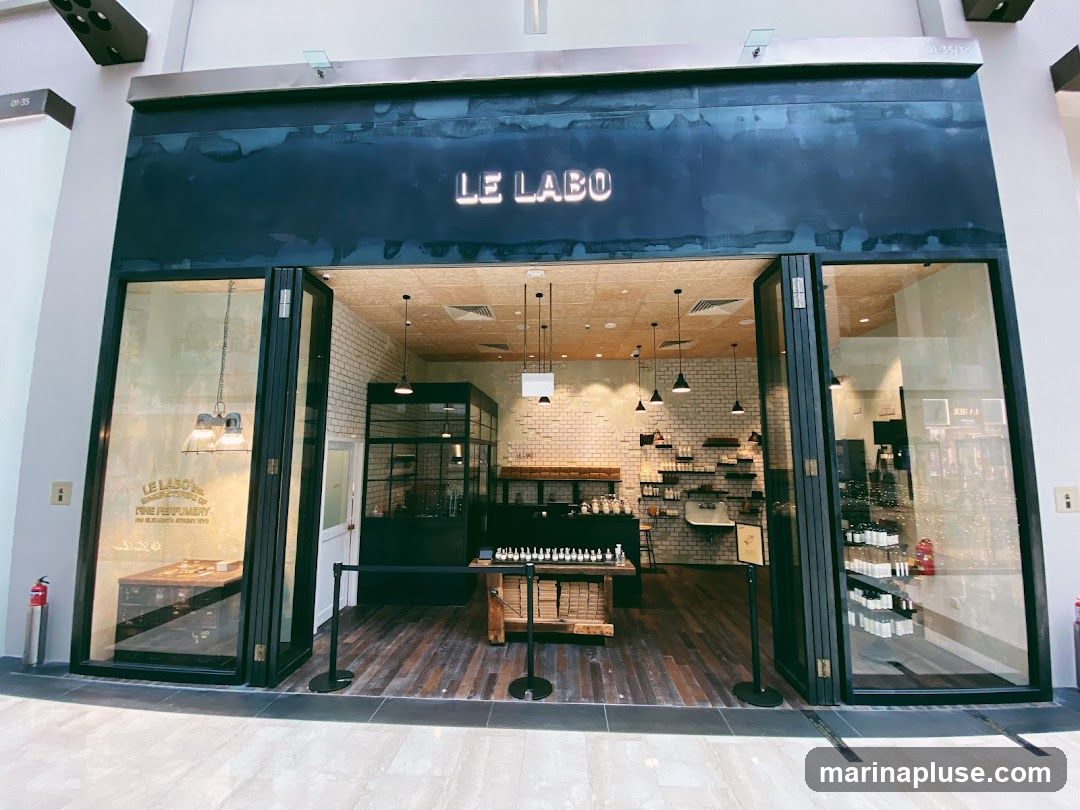 The exterior of a Le Labo store with open folding doors. The store has a rustic interior with exposed brick walls and wooden floors. Bottles of perfume are displayed on shelves and tables. The store's name, "LE LABO", is visible in large illuminated letters above the entrance. A sign on the left window reads "LE LABO FINE PERFUMERY".