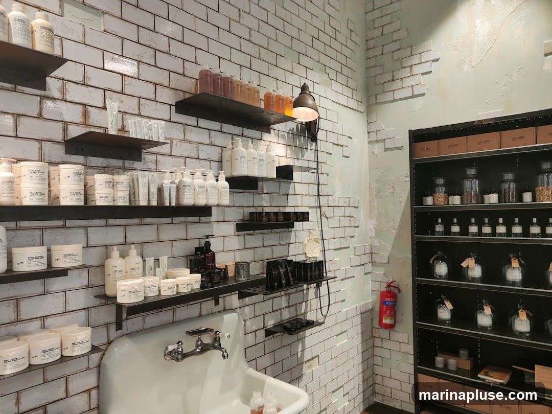 A display of various cosmetic products on shelves against a white brick wall. Products include bottles, jars, and tubes of skincare and haircare items. A sink is visible in the foreground. A tall black shelving unit on the right displays more products in jars and bottles, along with boxed items. A red fire extinguisher is mounted on the wall.