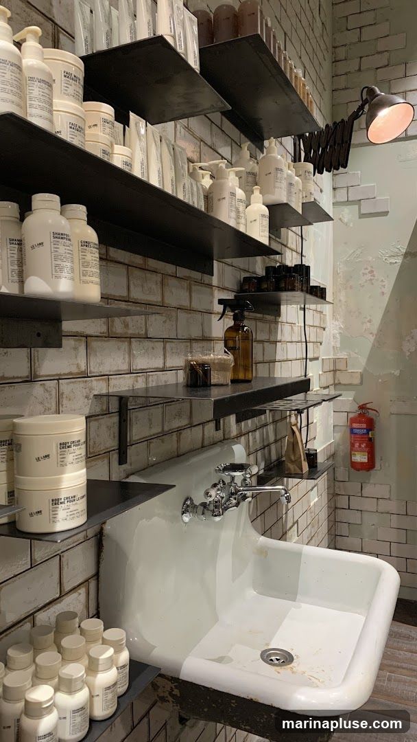 Shelves are stocked with various Le Labo branded products, including bottles, jars, and tubes, in white and amber packaging. A large white sink with chrome fixtures is in the foreground. A fire extinguisher is mounted on the wall. The walls are tiled with white subway tiles and some exposed brick.