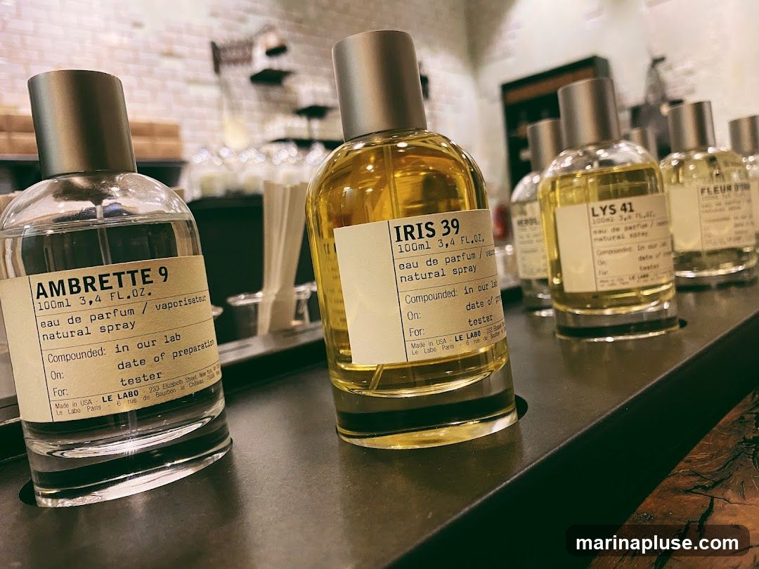 A close-up shot of several perfume bottles from the brand LE LABO, displayed on a dark surface. The bottles are clear glass with silver caps and feature cream-colored labels with handwritten-style text. Visible labels include 'AMBRETTE 9', 'IRIS 39', 'LYS 41', and 'FLEUR D'. The background is blurred, showing shelves with more items and a brick wall.
