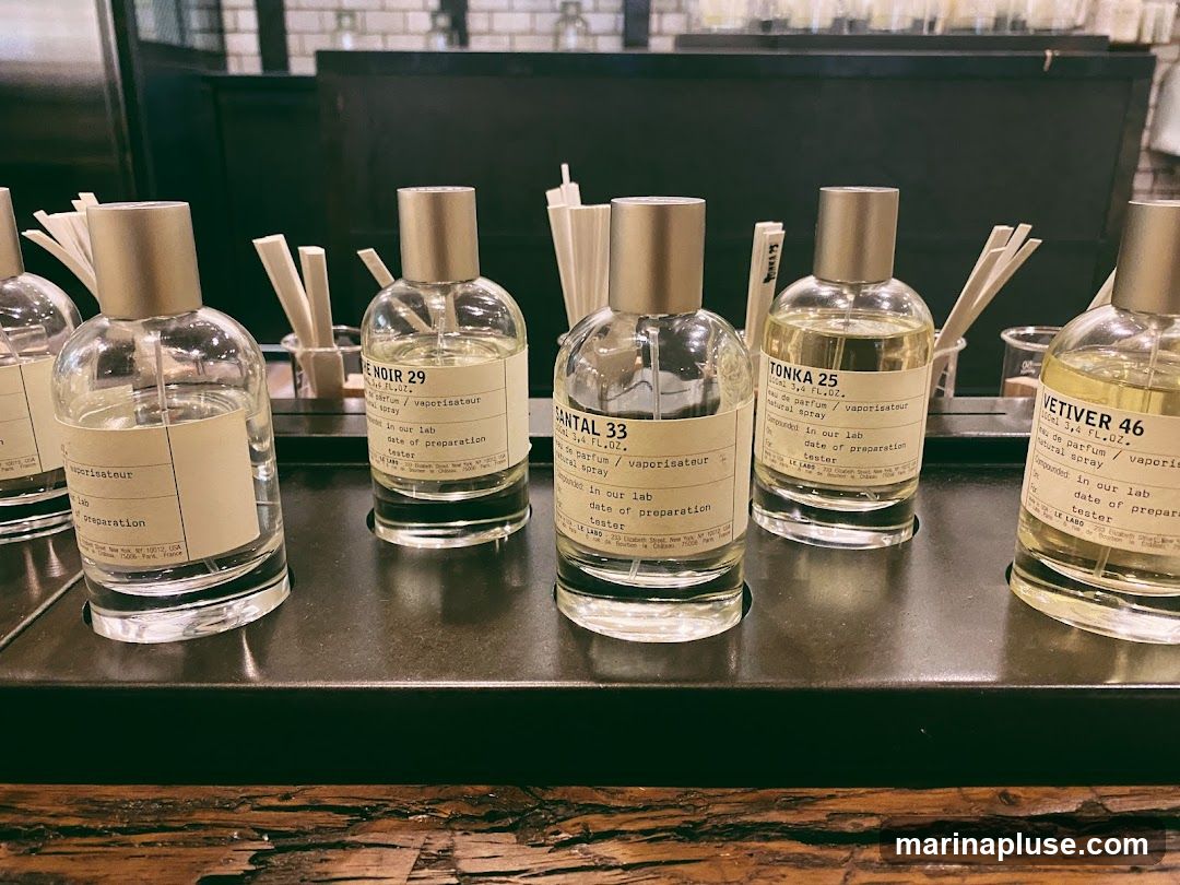 A close-up shot of several perfume bottles with labels, arranged on a dark surface. The labels indicate different scents like 'LE NOIR 29', 'SANTAL 33', 'TONKA 25', and 'VETIVER 46'. Each bottle is a clear glass spray bottle with a metallic cap and a white label detailing the scent name, size, and origin. Small paper scent strips are visible behind the bottles.
