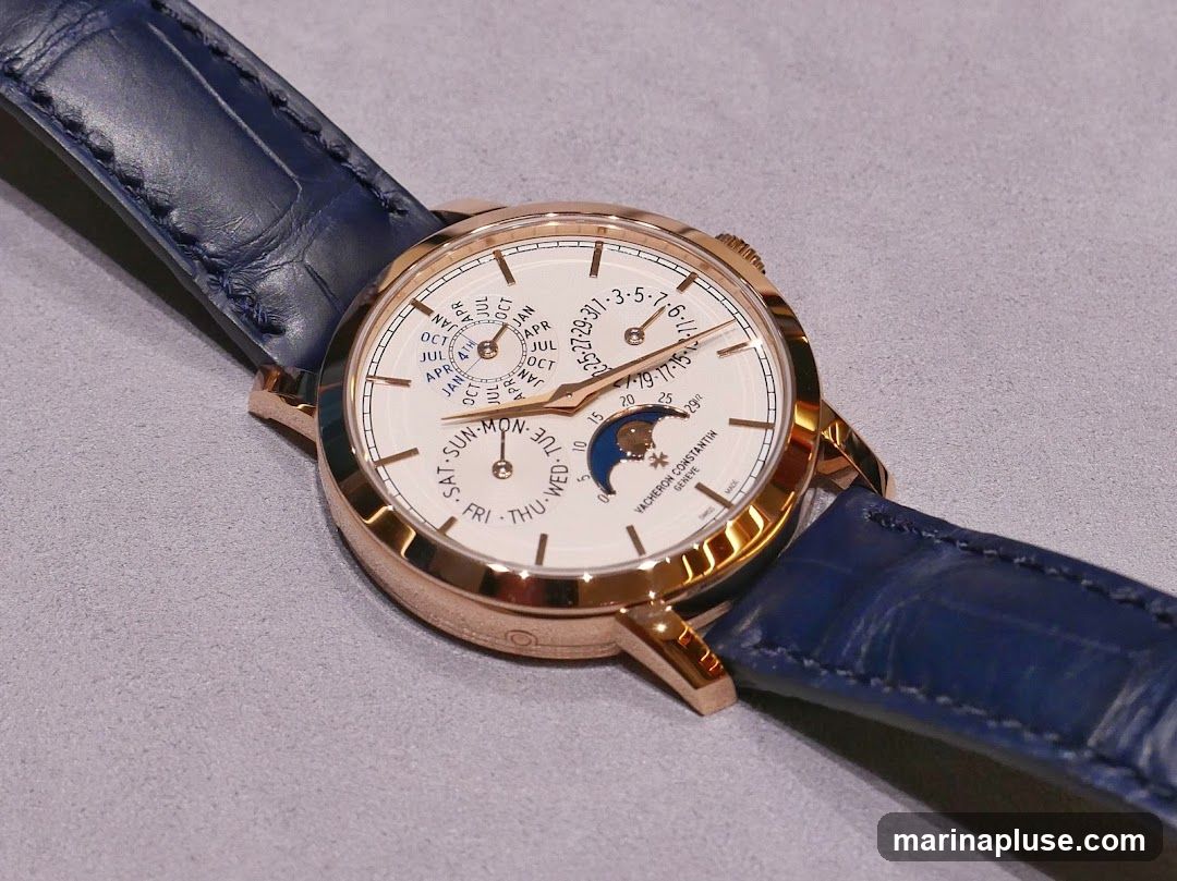A close-up shot of a Vacheron Constantin watch with a white dial, rose gold case, and blue alligator leather strap. The watch face displays multiple complications, including day, date, month, and moon phase indicators. The brand name "VACHERON CONSTANTIN GENEVE" is visible on the dial.