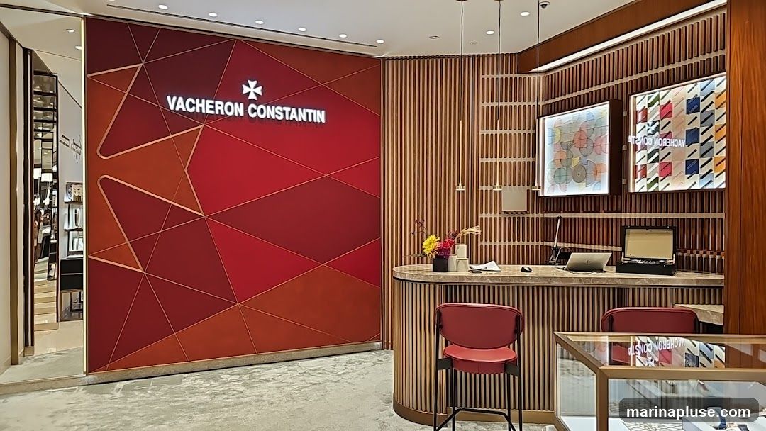 The interior of a Vacheron Constantin store features a large red wall with a geometric pattern and the brand's logo. A reception desk with two red chairs is visible, along with display cases and artwork on the wall.