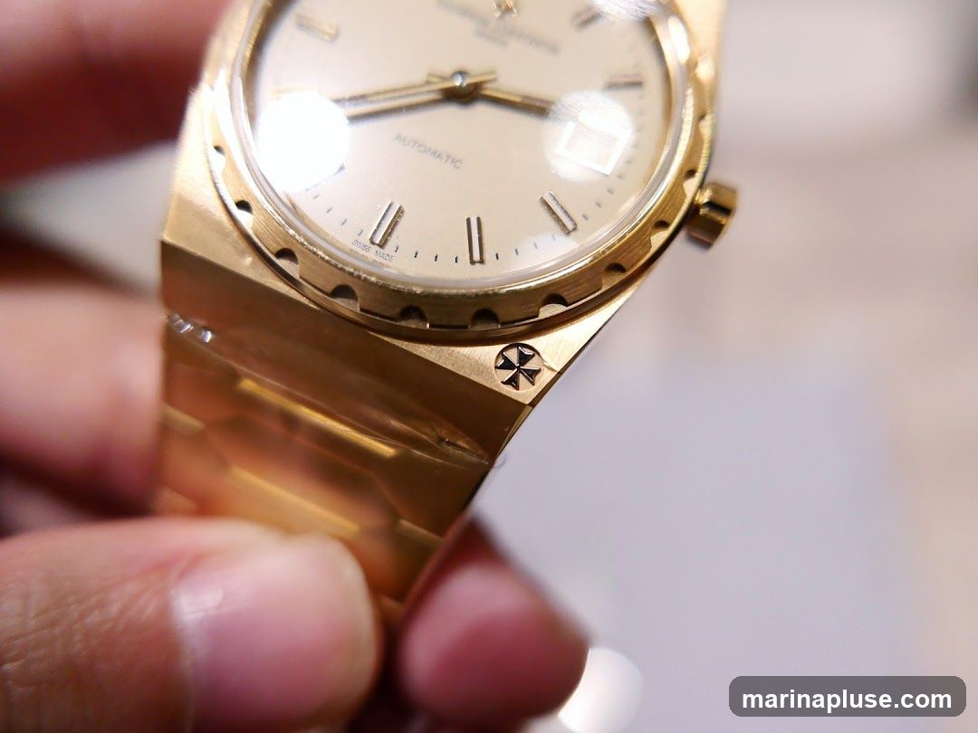 A close-up shot of a gold watch, held by a person's fingers. The watch face is light-colored with gold hands and hour markers. The word "AUTOMATIC" is visible on the face, along with "SWISS MADE". The watch band is also gold and appears to be made of interlocking links. A small, intricate cross-like emblem is visible on the side of the watch case.