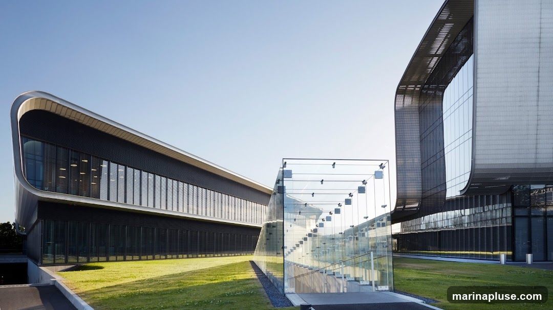 A wide shot of a contemporary office building with a curved facade and extensive glass elements. The building features a sleek, metallic exterior with horizontal window bands and blinds. A glass staircase enclosure is prominently visible, leading into the structure. The surrounding area is a well-maintained lawn with a clear blue sky overhead.