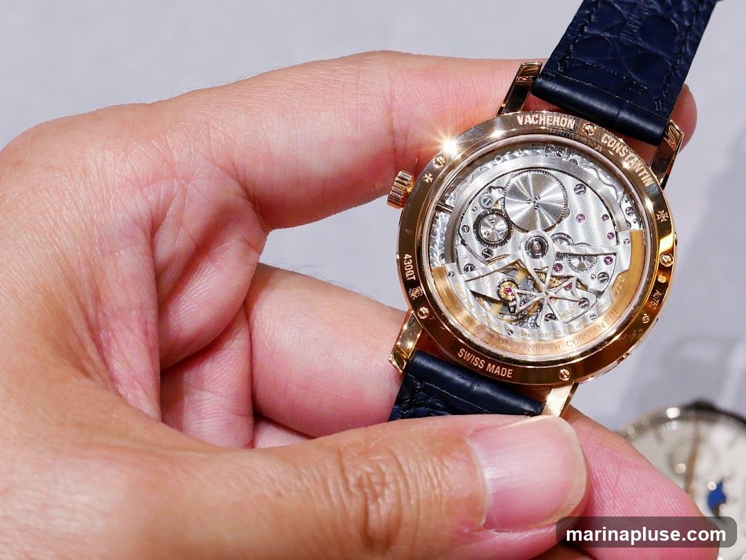 A close-up shot of the intricate mechanical movement of a Vacheron Constantin watch, held by a person's hand. The rose gold case and dark blue alligator strap are visible. The watch is engraved with "Vacheron Constantin," "Swiss Made," and other markings.