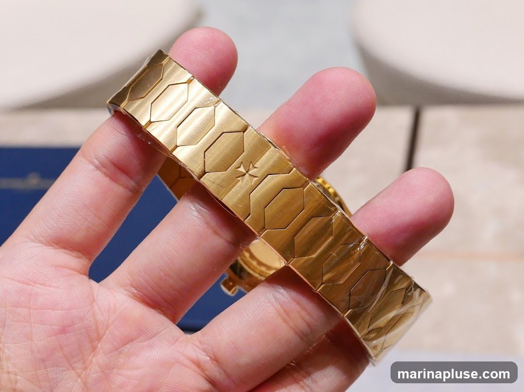 A close-up shot of a person's hand holding a gold watch bracelet. The bracelet has a unique geometric pattern with hexagonal links and a star-shaped emblem in the center. The watch face is partially visible behind the bracelet. The background is blurred, showing hints of furniture and a blue box.