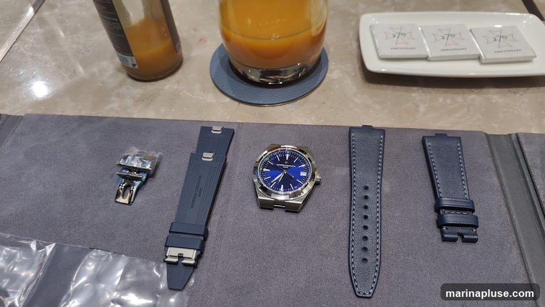 A blue dial watch with silver casing is displayed on a grey surface. Next to it are two blue watch straps and a watch buckle. In the background, a glass of orange juice and a bottle are visible, along with a white plate holding three small wrapped items labeled "270 ANNIVERSARY".