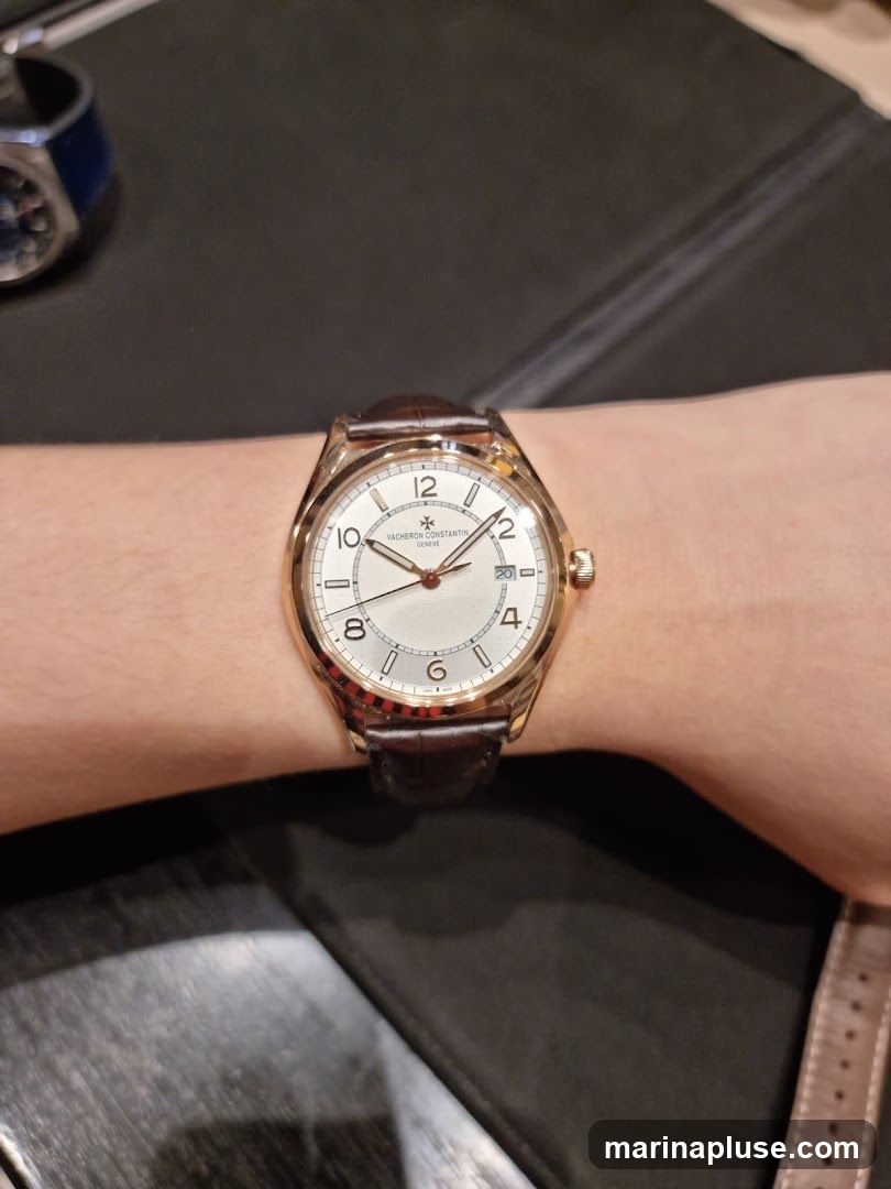 A close-up shot of a Vacheron Constantin watch on a person's wrist. The watch has a white dial with rose gold casing and a brown leather strap. The dial features Arabic numerals for hours 12, 2, 4, 6, 8, 10, and stick markers for the other hours. The brand name "VACHERON CONSTANTIN GENEVE" is visible on the dial. A date window is present at the 3 o'clock position. In the background, another watch with a blue strap is partially visible.