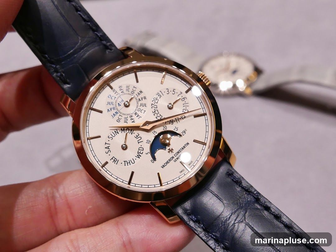 A close-up shot of a Vacheron Constantin watch with a rose gold case and a dark blue alligator leather strap. The watch face displays multiple complications, including a moon phase indicator, day of the week, date, and month. Another watch with a white face and a light-colored strap is visible in the blurred background.