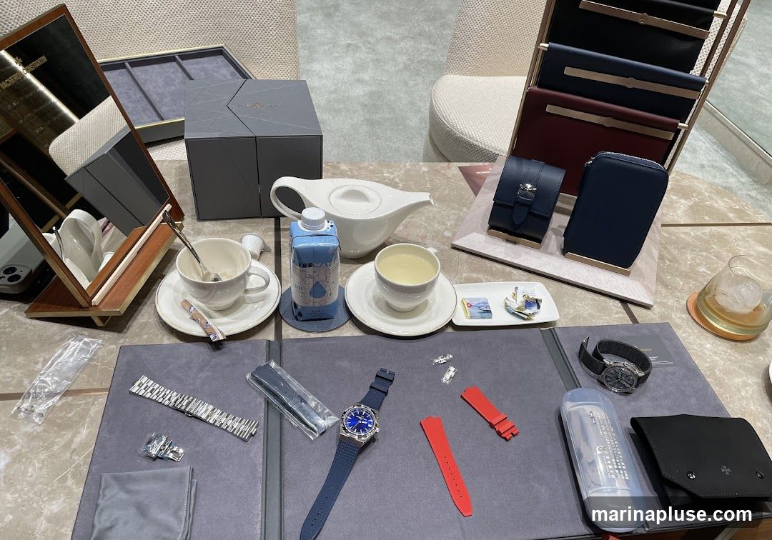 A table is set with luxury watches, watch straps, and accessories, alongside refreshments. A white teapot, a cup of tea, and a bottle of water are arranged on the table. Several watches, including one with a blue strap and another with a black strap, are displayed. Various watch straps, a metal watch band, and cleaning cloths are also visible. In the background, a display stand holds several leather pouches or wallets in dark colors. A mirror reflects the Vacheron Constantin logo.