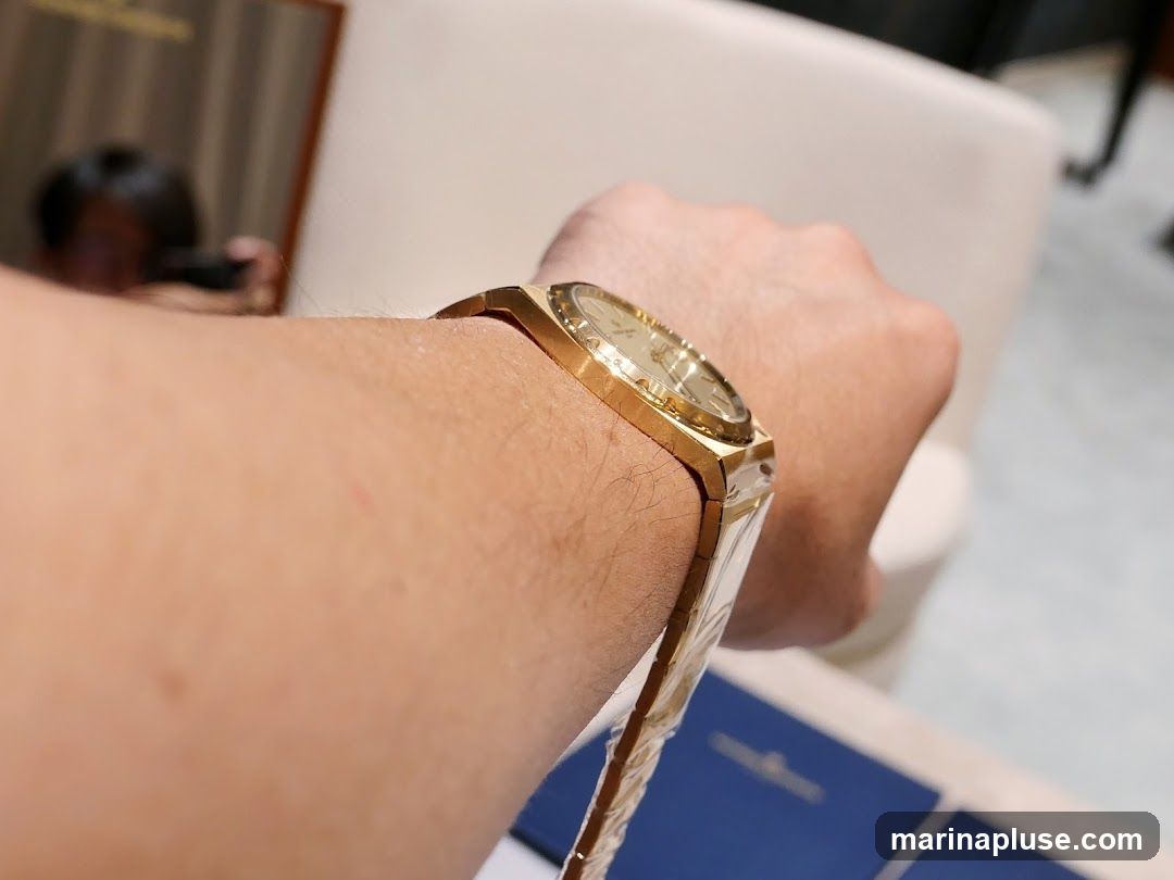 A close-up shot of a person's wrist wearing a gold-toned watch. The watch has a rectangular case and a metallic band. In the background, a blurred reflection of a person holding a camera can be seen in a mirror.