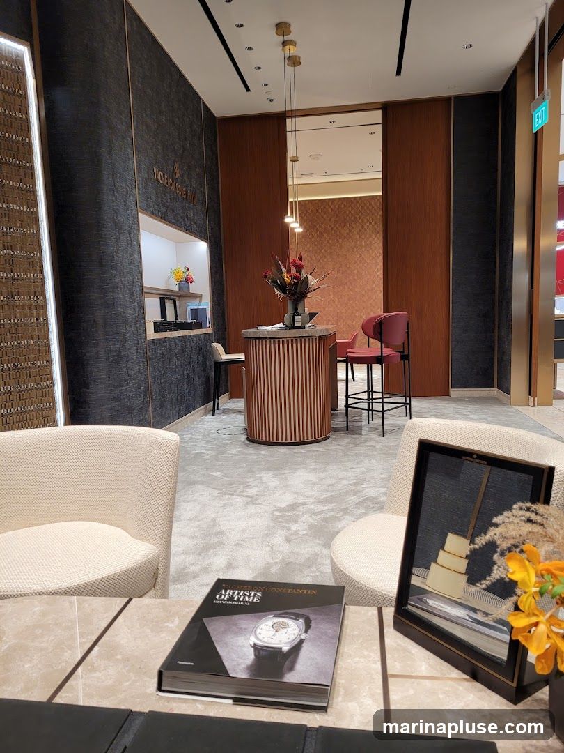 The interior of a high-end watch store, featuring plush seating areas, a central display counter with floral arrangements, and elegant wall paneling. A book about Vacheron Constantin watches is prominently displayed on a marble-topped table.