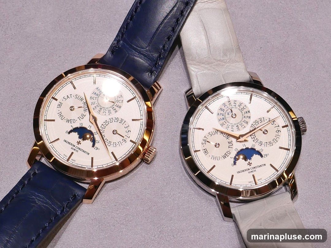 Two luxury Vacheron Constantin watches with white dials and leather straps are displayed. The watch on the left has a rose gold case and a dark blue strap, while the watch on the right has a silver-toned case and a light grey strap. Both watches feature complex dials with day, date, month, and moon phase complications. The brand name "VACHERON CONSTANTIN GENEVE" is visible on both dials.