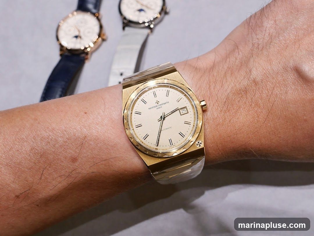 A close-up shot of a person's wrist wearing a gold watch with a cream-colored face. The watch has Roman numerals and a date window. In the blurred background, two other watches are visible, one with a blue strap and the other with a white strap.