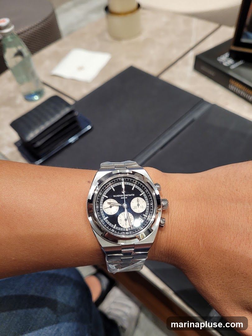 A close-up shot of a Vacheron Constantin watch on a person's wrist. The watch has a dark blue dial with silver hands and markers, and three subdials. The watch is on a stainless steel bracelet. In the background, there is a table with a bottle of water, a wallet, and a book.