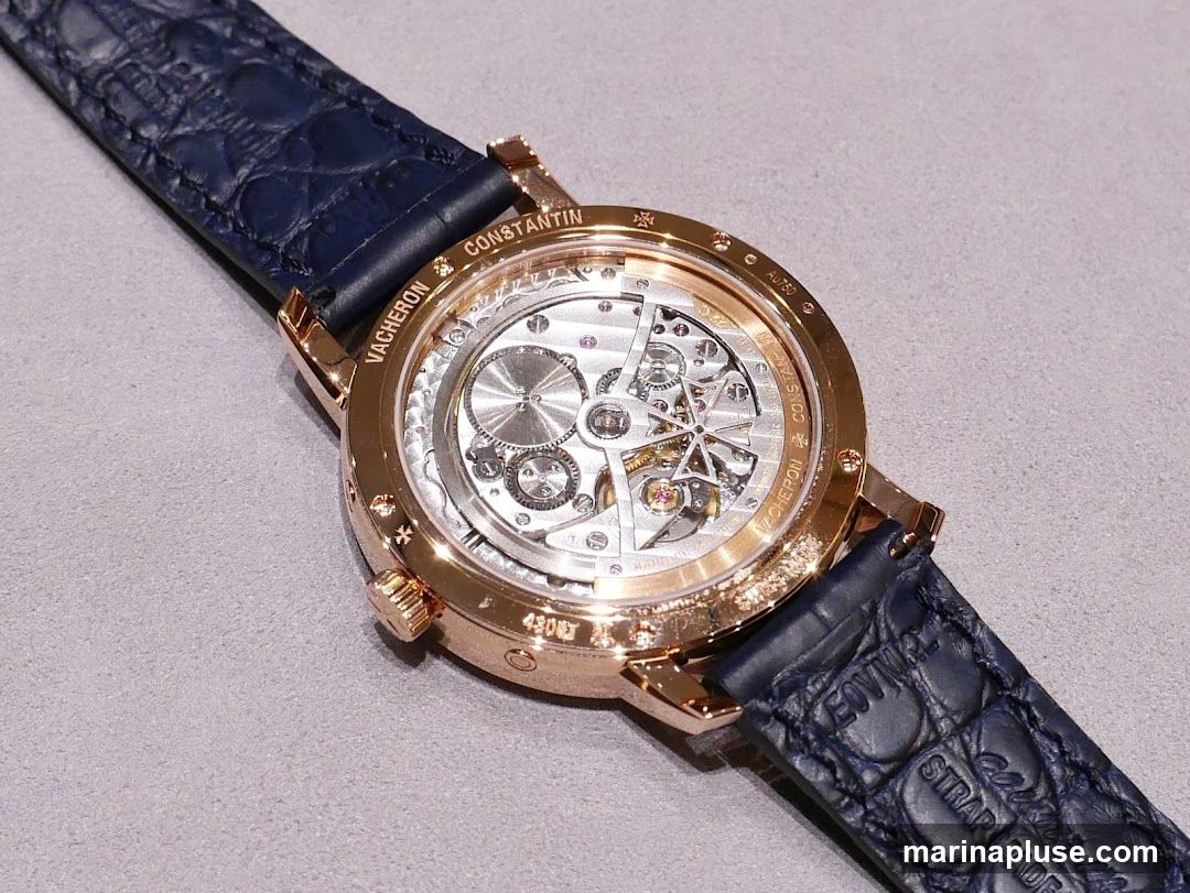 A close-up view of the intricate mechanical movement of a Vacheron Constantin watch, seen through the transparent case back. The rose gold case is visible, along with a dark blue alligator leather strap. Engravings on the case include 'VACHERON CONSTANTIN' and 'AU750'.