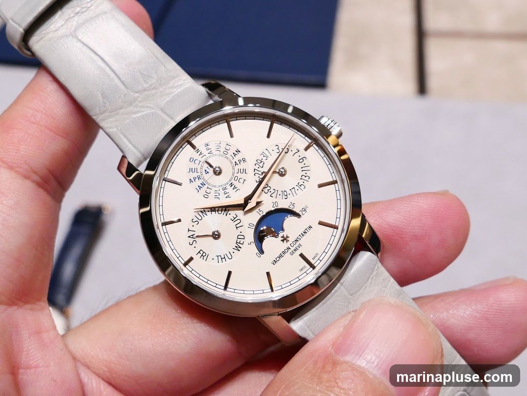 A close-up shot of a Vacheron Constantin watch with a light-colored alligator strap. The watch face is cream-colored with multiple complications, including a date display, day of the week, month, and moon phase. The brand name and "GENEVE" are visible on the lower part of the watch face.