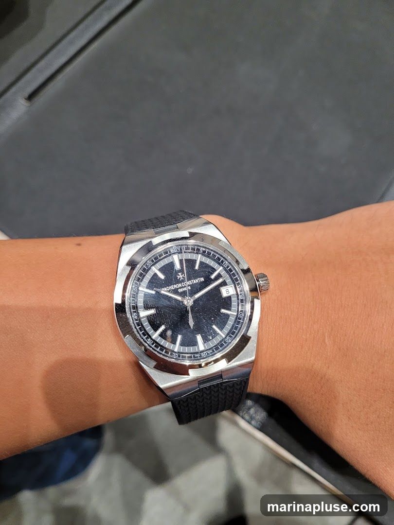 A close-up shot of a Vacheron Constantin watch on a person's wrist. The watch has a black dial with silver hands and hour markers. The date is visible at the 3 o'clock position. The watch has a stainless steel case and a black rubber strap. The background is dark and out of focus.